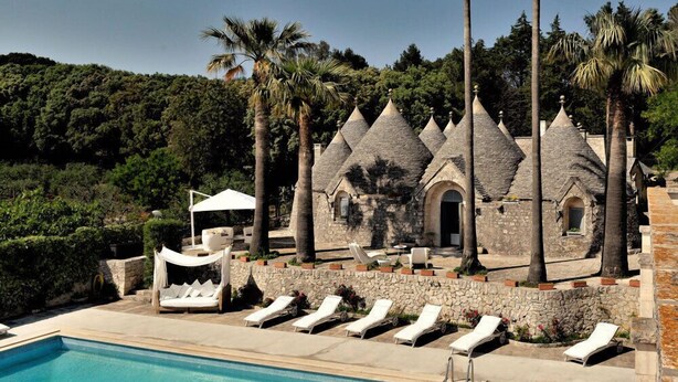Ein Poolbereich mit Liegestühlen, ein Steinhaus mit Kegeldächern und Palmen.