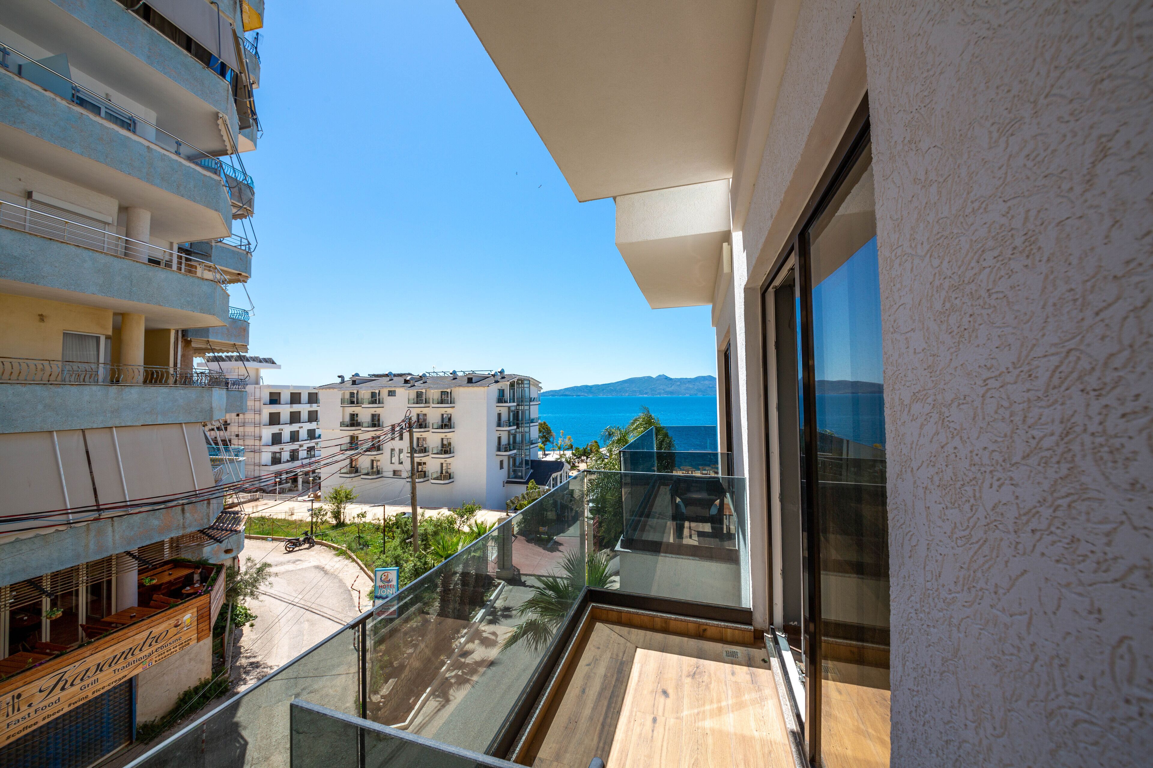 A balcony with a clear view of the sea and mountains, adjacent buildings, and a restaurant with outdoor seating.