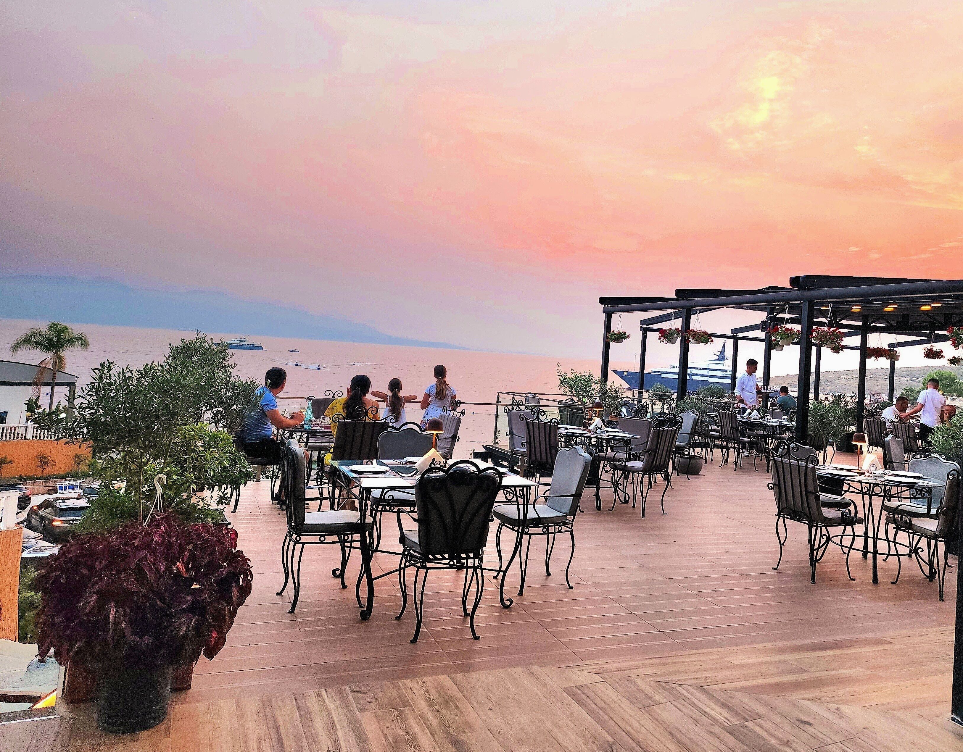 Een terras met tafels en stoelen, met uitzicht op een waterpartij en een zonsondergang.