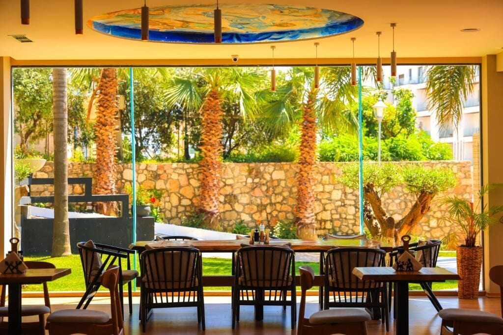 Un comedor interior con vista a una pared de piedra y palmeras. Mesas y sillas dispuestas para comer.