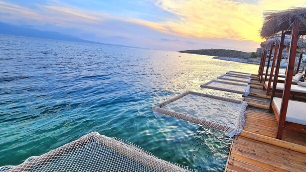 A wooden deck with lounge chairs and a thatched roof structure overlooking a body of water. - Hotel Apollon Saranda (Sarandë)