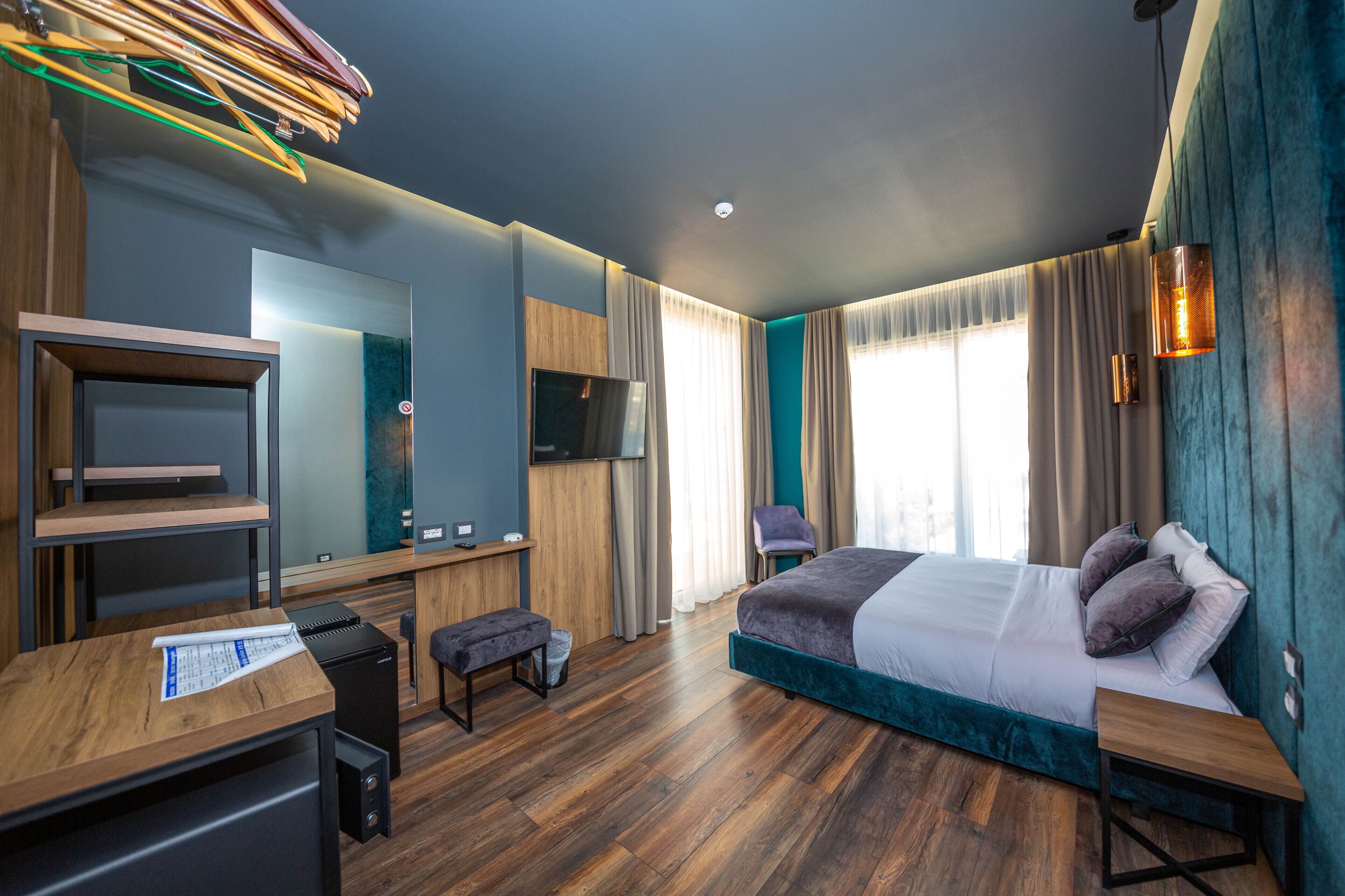 Una habitación de hotel moderna con un escritorio de madera, una cama con cabecera azul, un televisor y un ventanal grande con cortinas.
