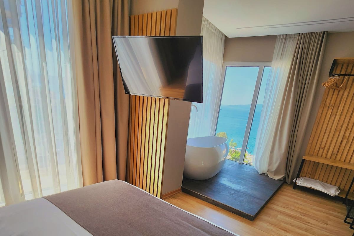 A hotel room with a large bed, a flat-screen TV, a view of the sea, and a wooden floor.