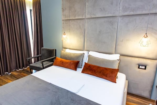 A modern hotel room with a large bed, two bedside tables, a chair, and a headboard with built-in lighting. - Hotel Apollon Saranda (Sarandë)