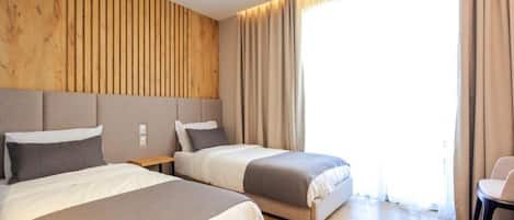 Habitación de hotel con dos camas, cabecera de madera, un ventanal grande con cortinas y una mesita con una silla.