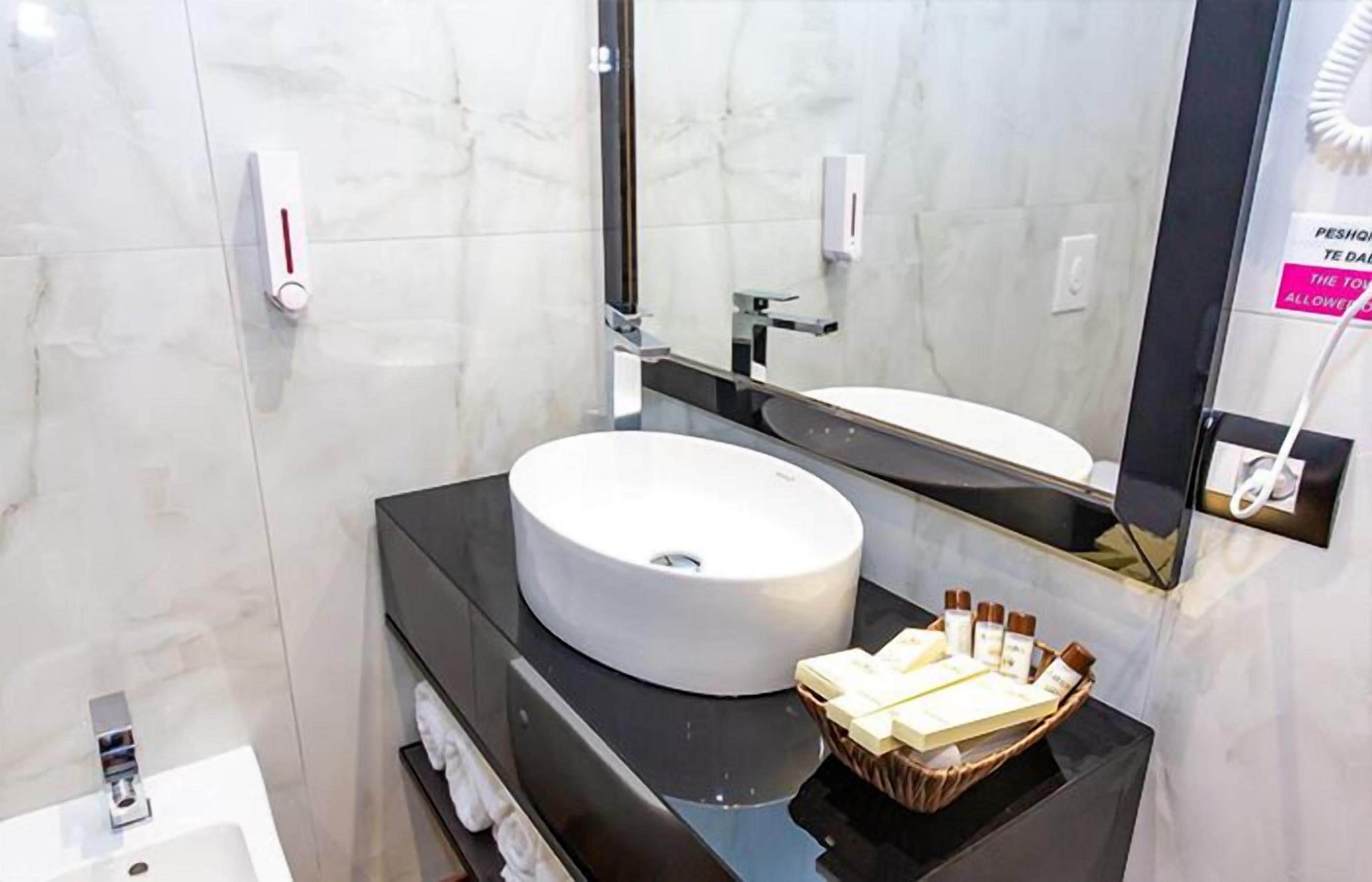 A modern bathroom with a white sink, black countertop, and a mirror. There is a towel rack and a basket of toiletries.