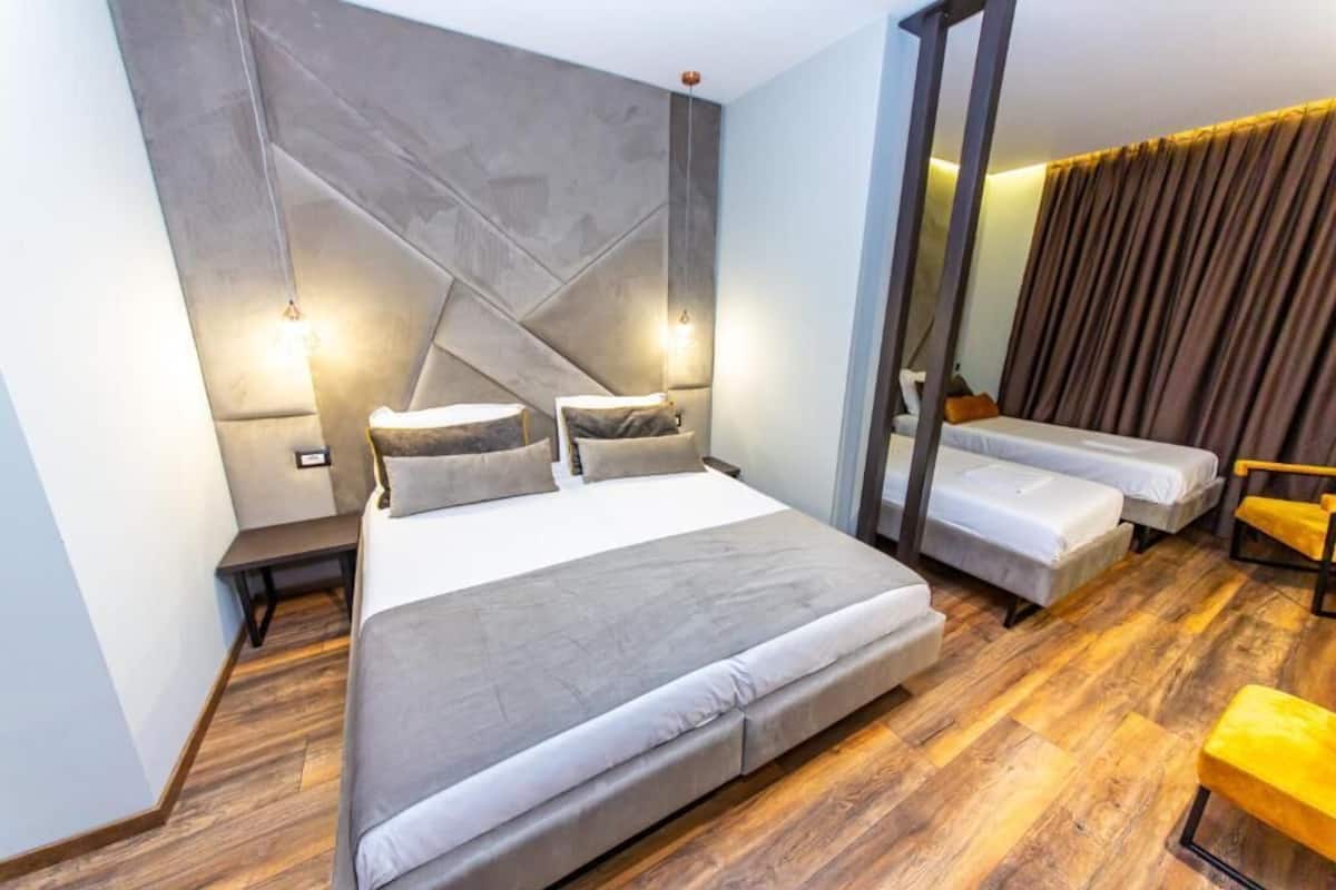 A modern hotel room with a large bed, two smaller beds, a wooden floor, and a mirror on the wall.