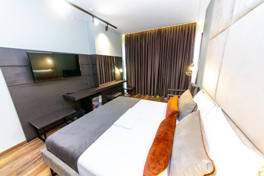 A modern hotel room with a flat-screen TV mounted on the wall, a bed with white and brown bedding, a desk with a chair, and a large window with curtains.