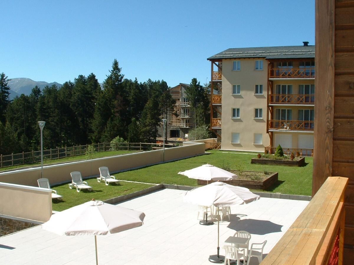 A rooftop terrace with white lounge chairs and umbrellas, a pool, and a view of a forest and buildings in the background.