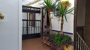 Un estrecho pasillo exterior con suelo de baldosas, una reja negra y un pequeño jardín con plantas y una jardinera de madera.