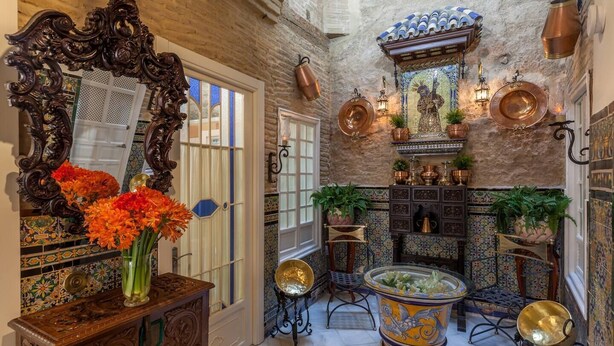 Una habitación con un espejo grande y ornamentado, un aparador de madera con un florero, una mesa decorativa con un cuenco y una pared con ollas de cobre y un mural de azulejos.