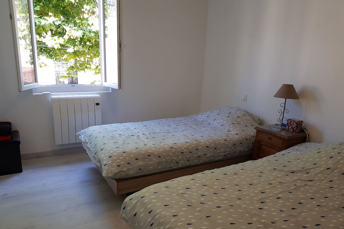 A bedroom with two beds, a window with a view of trees, a radiator, and a bedside table with a lamp.