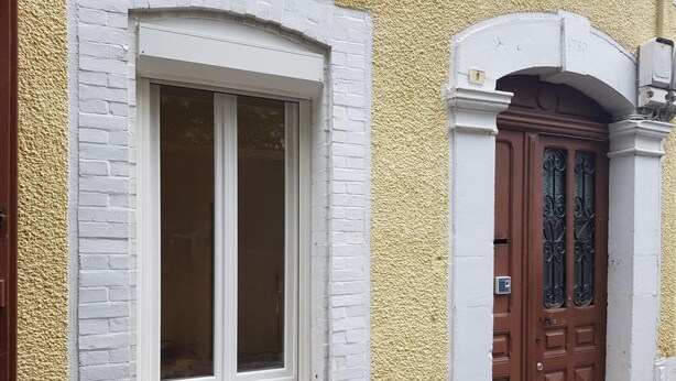 A building with a white brick facade, a window with white trim, and a brown door with decorative ironwork.