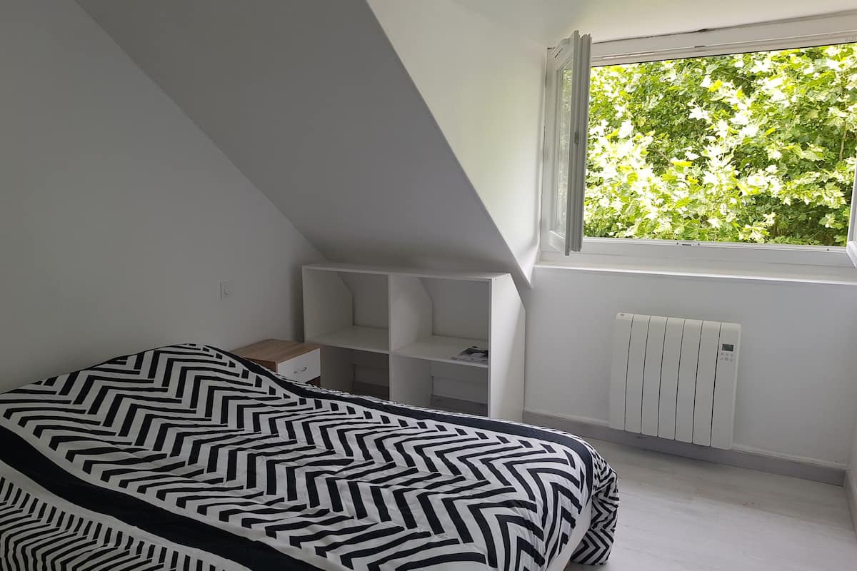 A bedroom with a bed, a window with a view of greenery, a radiator, and built-in shelving.