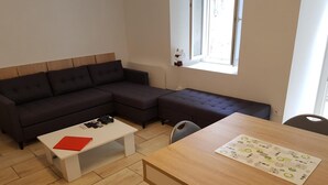 A modern living room with a dark sectional sofa, a white coffee table with remotes, and a wooden dining table with a placemat.