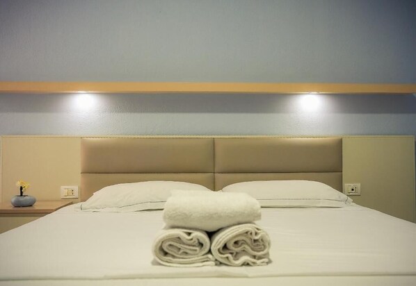 A neatly made bed with a folded towel in the center, a bedside table with a vase of flowers, and a wall-mounted light fixture above the bed.