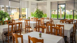 A restaurant with multiple tables and chairs, large windows with blinds, and indoor plants. - Hotel Andi (Rrashbull)