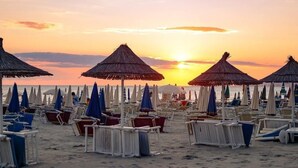 A beach with rows of sun loungers and umbrellas, a clear sky, and the sun setting in the background. - Hotel Andi (Rrashbull)