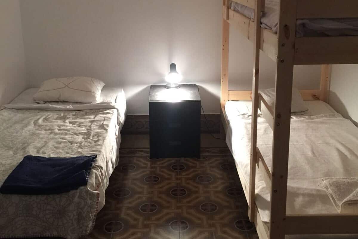Una habitación pequeña y tenuemente iluminada con dos camas individuales y una litera, una mesita con una lámpara, y baldosas de suelo estampadas.