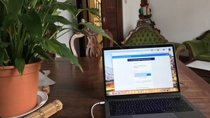 Un ordenador portátil sobre una mesa de madera, con una planta en maceta, una silla de madera, y una estantería en una habitación con una puerta de madera y un sillón tapizado de color verde.