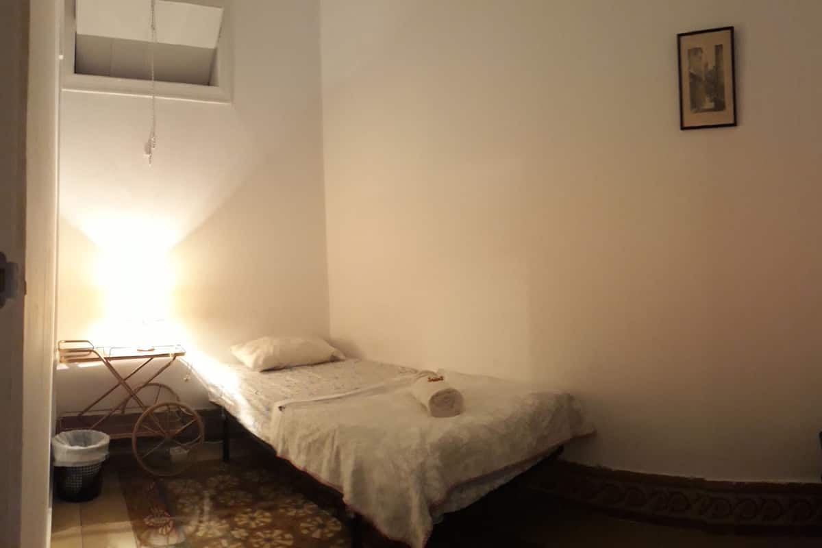 Una habitación pequeña y poco iluminada, con una cama individual, una mesita con una silla y una ventana con persianas.