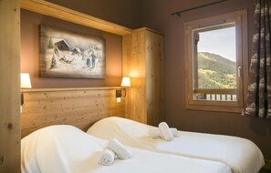 Une chambre d’hôtel avec deux lits, une tête de lit en bois, un tableau représentant un paysage enneigé, une fenêtre donnant sur des collines et deux lampes fixées au mur.