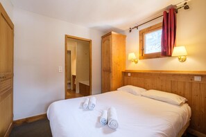 A hotel room with a wooden headboard, a bed with white linens, two rolled towels on the bed, a wooden wardrobe, a bathroom visible through an open door, and two wall-mounted lamps. - 200m from resort center, fitness, swimming pool, sauna, hammam, balcony, tv, ski locker, 72m² (Lanslevillard)