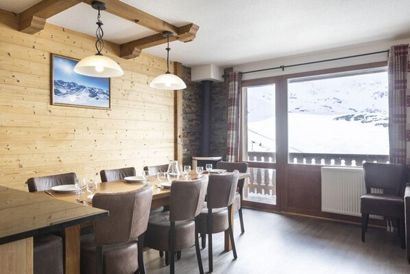 A dining area with wooden walls, a large window with a view of snow-covered mountains, and a fireplace.