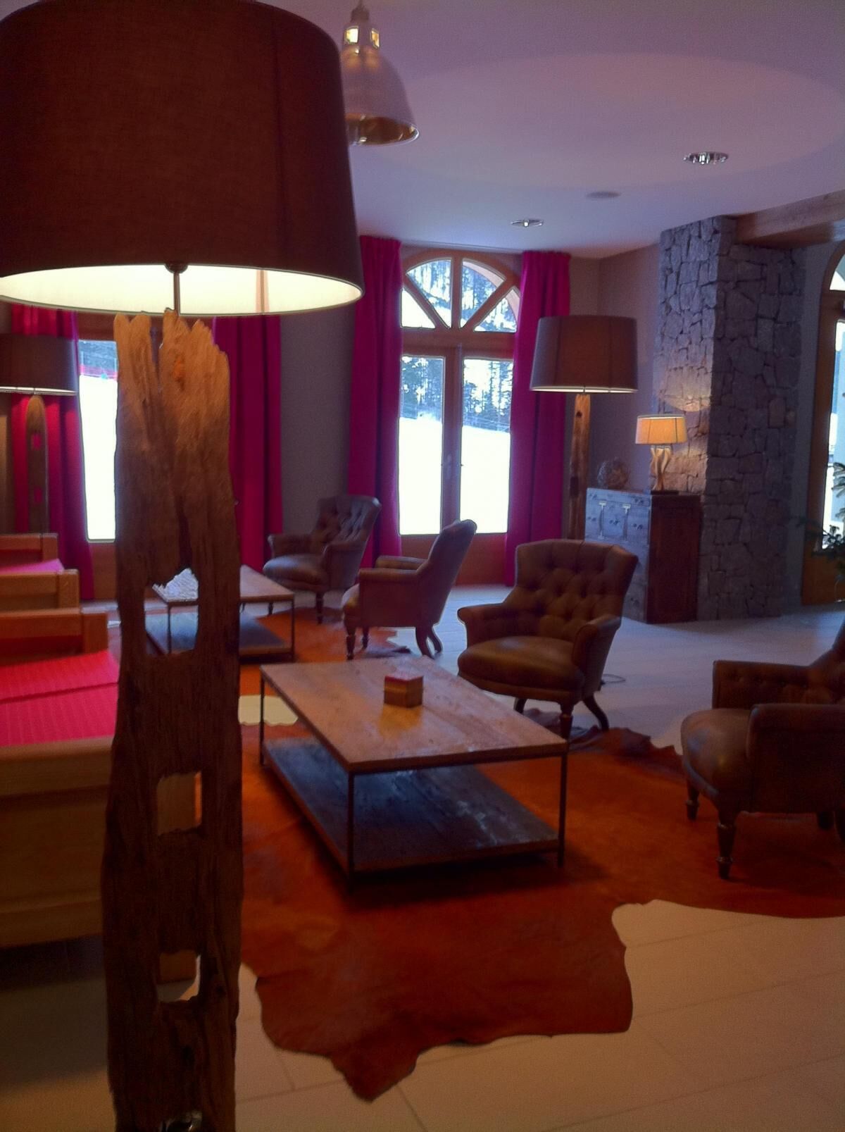 A spacious living room with a stone wall, leather armchairs, a wooden coffee table, and a large window with curtains.