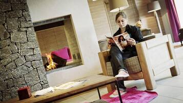 A person sitting on a wooden chair reading a magazine in a room with a stone wall, a fireplace, and a pink rug.