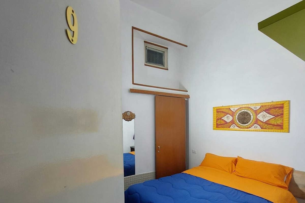 A bedroom with a bed, a door, a window, and a wall-mounted artwork.