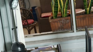 A room with a glass table, a black object on the table, and a window with a view of potted plants and furniture. - B&B Cappotto Civico 48 (Pompei)