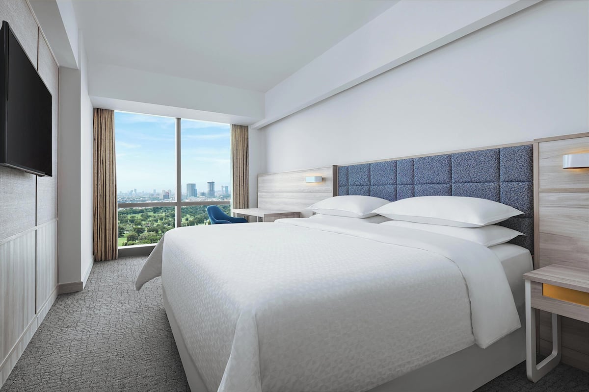 Ein modernes Hotelzimmer mit einem großen Bett, einem Flachbildfernseher, Blick auf die Stadt und einem kleinen Schreibtisch mit Lampe.