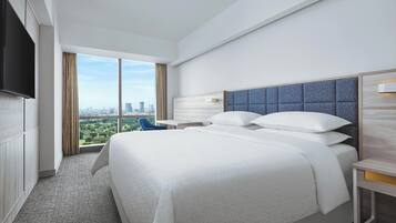 A modern hotel room with a large bed, a flat-screen TV, a city view, and a small desk with a lamp.