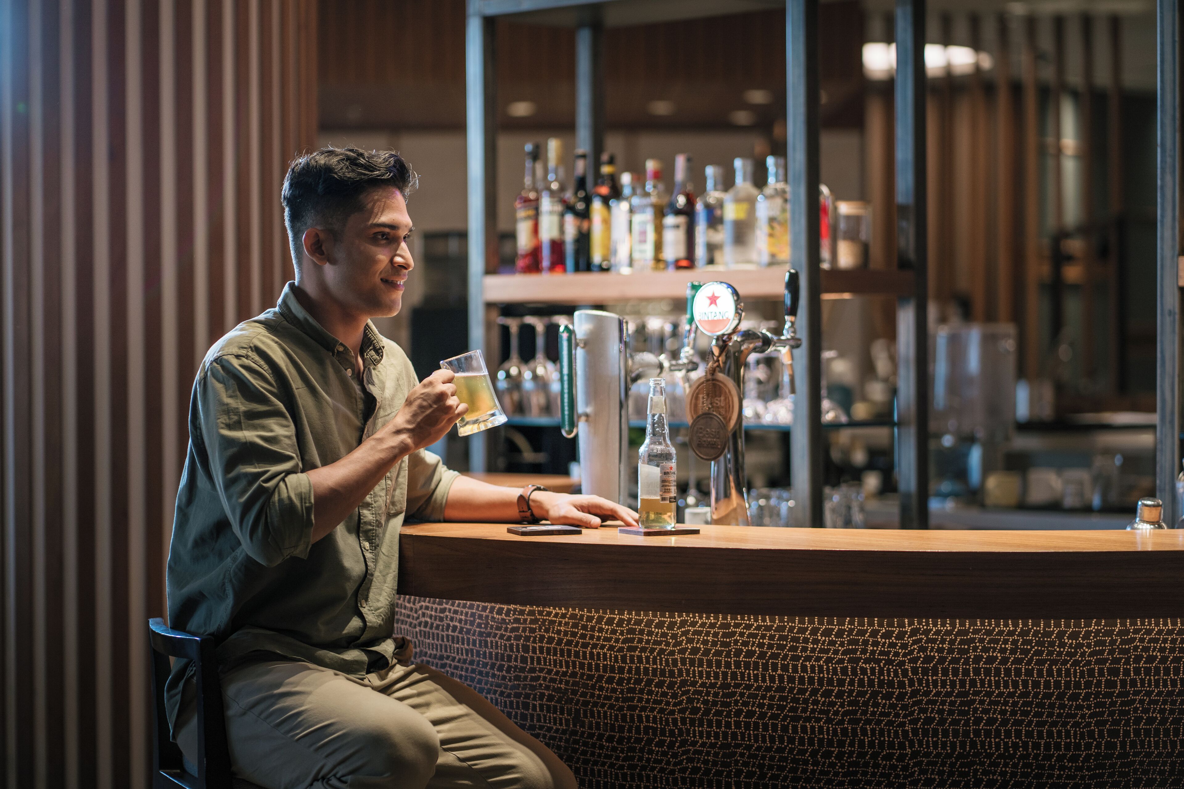 A person seated at a bar counter with a drink in hand, a tap, and various bottles in the background.