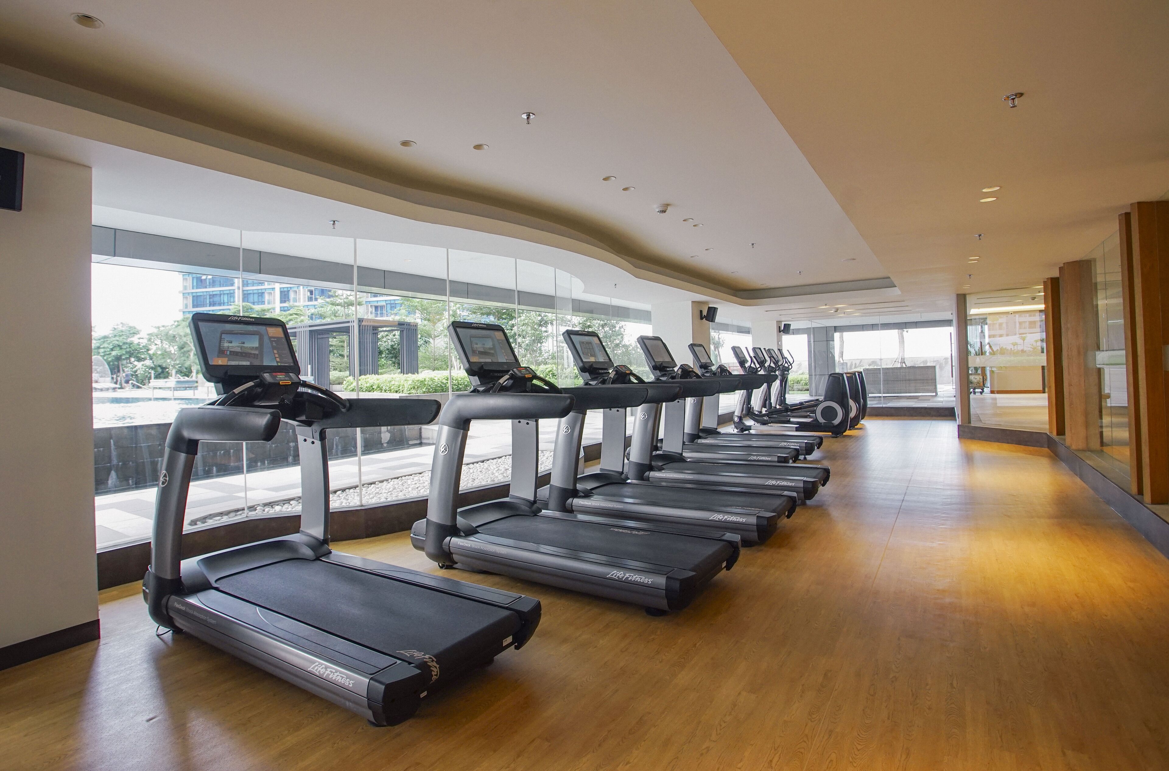 Une rangée de tapis de course dans une salle de sport dotée de grandes fenêtres et d’un plancher en bois.