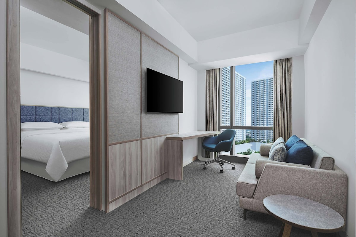 Ein modernes Hotelzimmer mit Bett, Schreibtisch, Fernseher, Sofa und Blick auf hohe Gebäude.