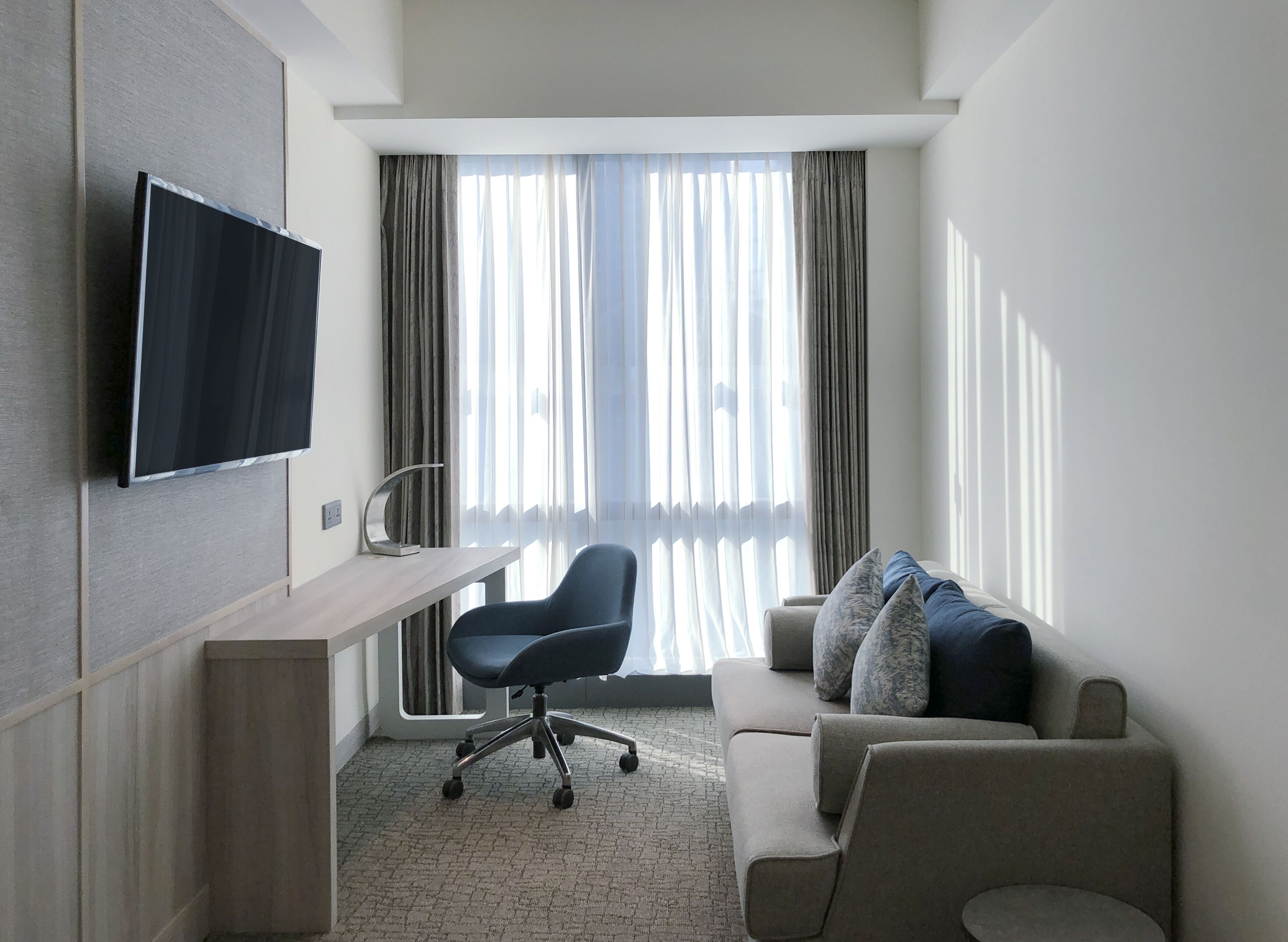 A modern hotel room with a flat-screen TV mounted on the wall, a desk with a chair, a sofa with cushions, and large windows with sheer curtains.