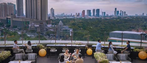 A rooftop dining area with outdoor seating, city skyline views, and potted plants.