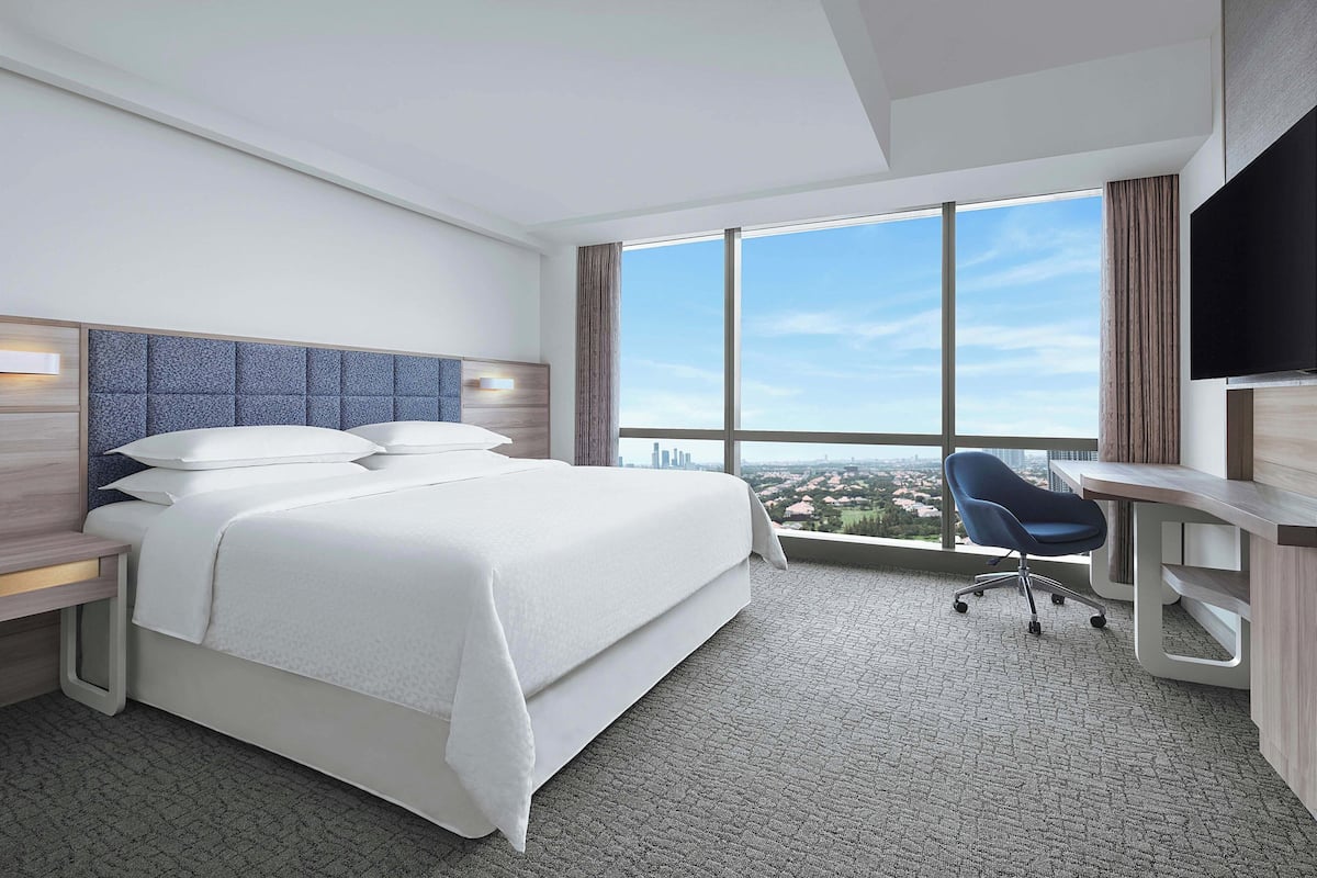 Ein Hotelzimmer mit einem großen Bett, einem Schreibtisch, einem Sessel, einem Fernseher und Blick auf die Stadt.