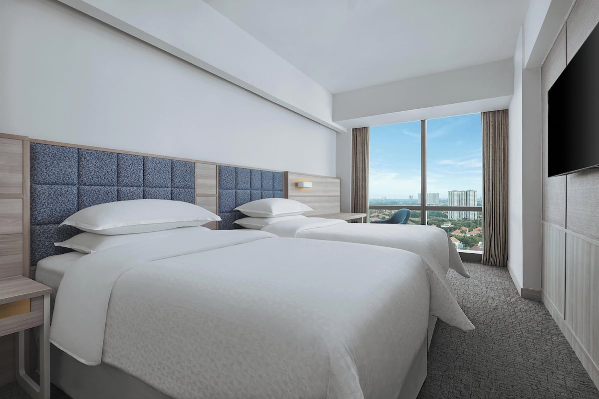 Ein Hotelzimmer mit zwei Betten, einem großen Fenster mit Blick auf die Stadt, einem Flachbildfernseher und einem Nachttisch.
