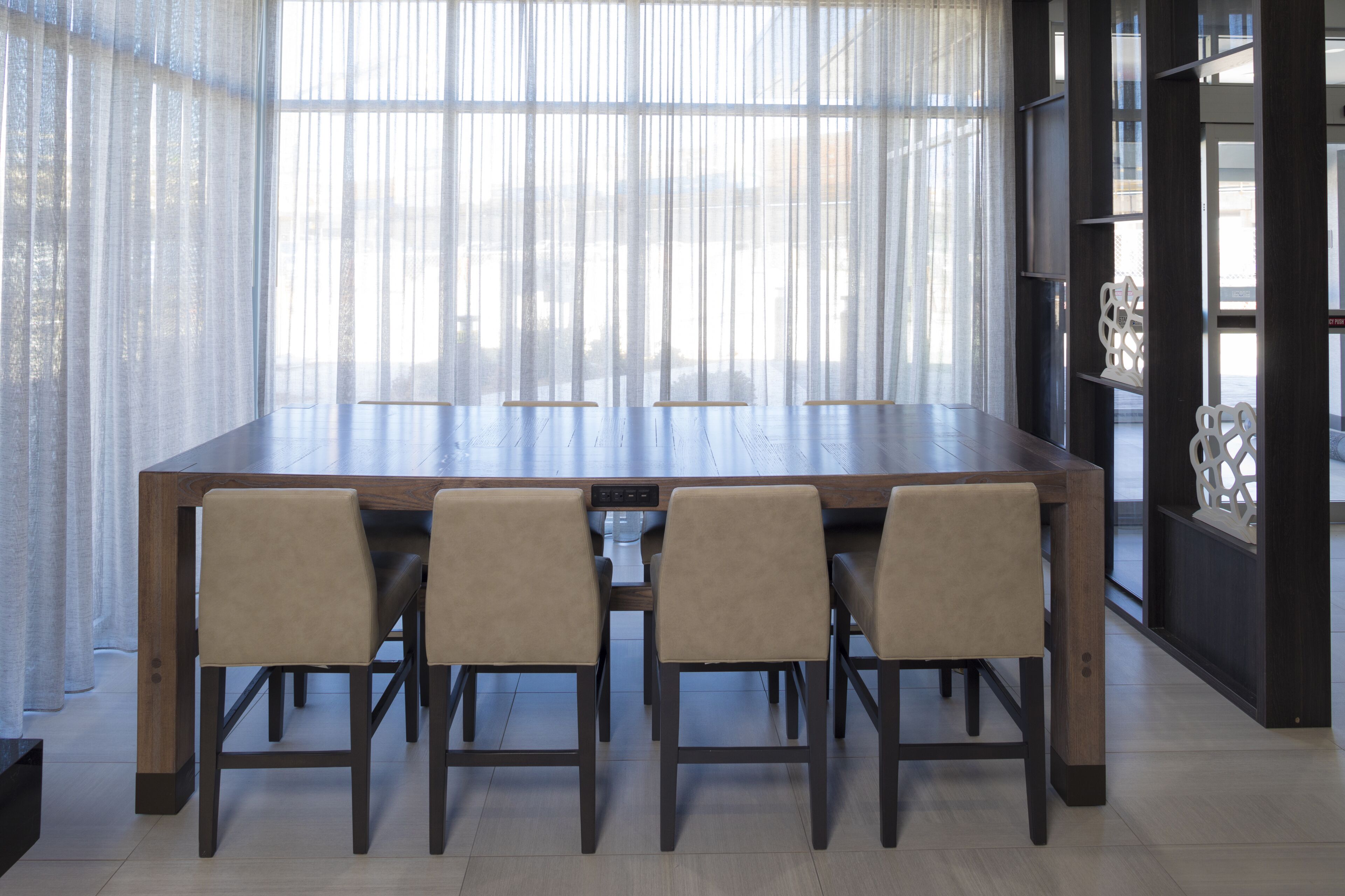 A dining area with a long wooden table and four chairs, large windows with sheer curtains, and a dark wooden bookshelf with decorative items.