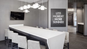A modern dining area with a long table, multiple chairs, pendant lighting, a flat-screen TV, and a motivational quote on the wall.