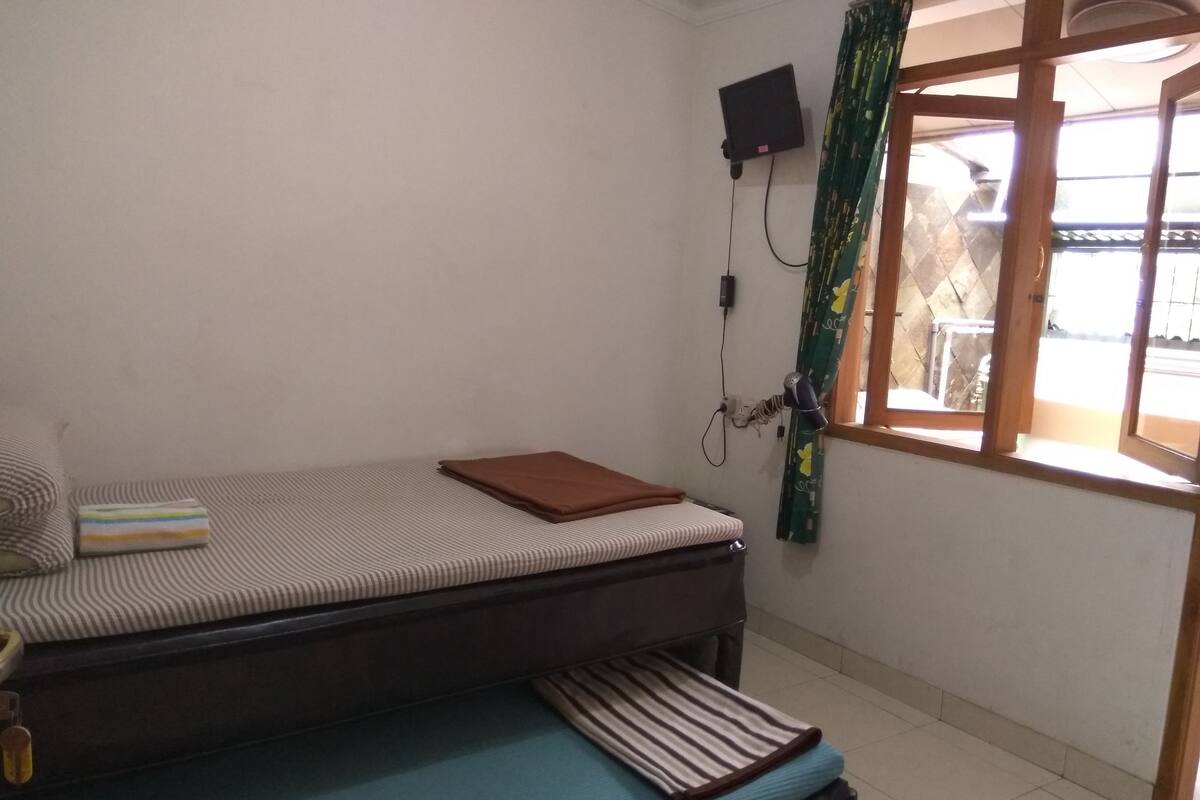 A bedroom with a single bed, a bedside table, a small table with a book, a wall-mounted TV, and a window with curtains.