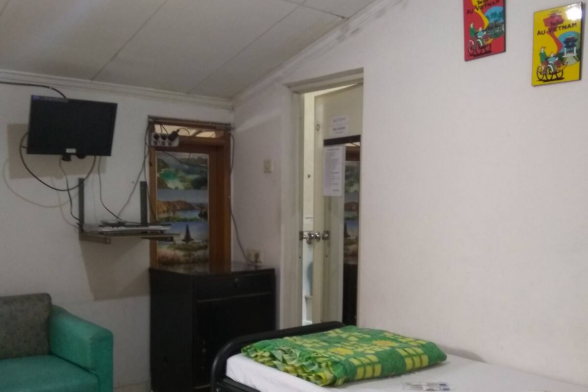 A small, compact room with a bed, a green sofa, a desk, and a television mounted on the wall.
