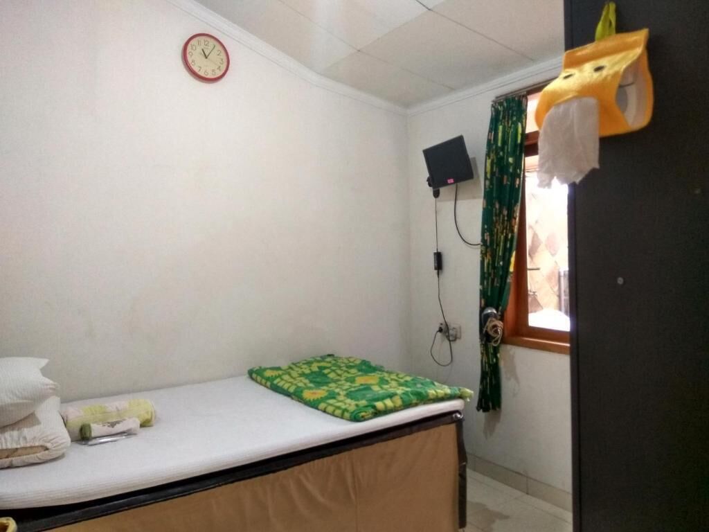 A small, simple room with a single bed, a clock on the wall, a window with curtains, and a mounted television.