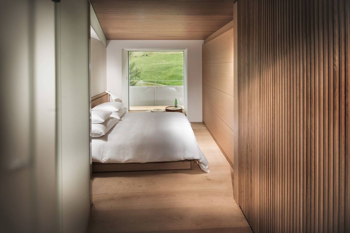 A narrow, single-bed room with a wooden floor, a large window with a view of a green landscape, and a bedside table with a lamp.