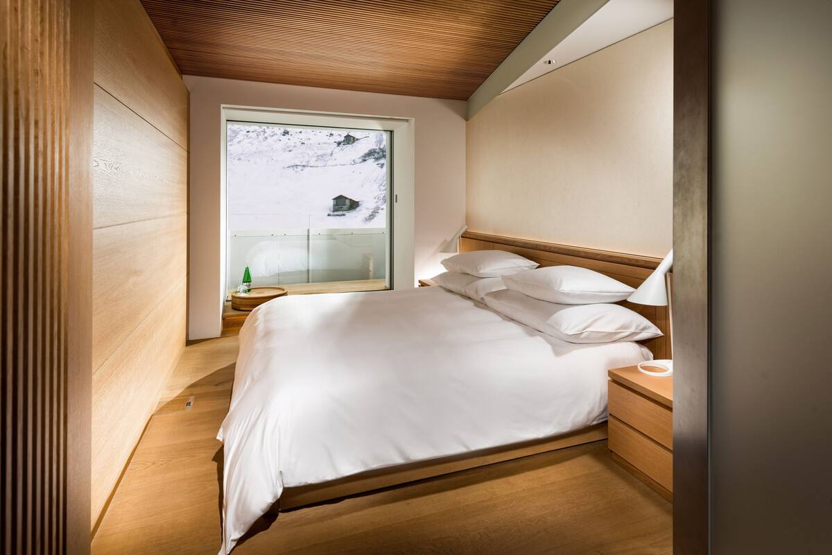 A bedroom with a wooden bed, bedside tables, a nightstand, and a window with a view of a snowy landscape.
