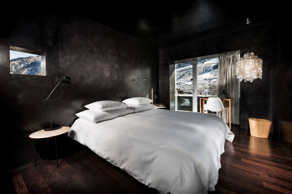 A modern bedroom with a large bed, a side table, a lamp, a view of a snowy landscape through a window, and a chandelier.