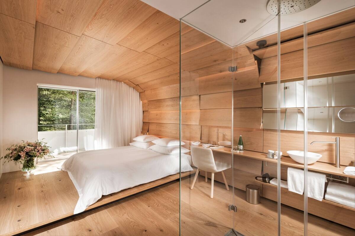 A modern bedroom with a wooden ceiling, a large bed, a desk, and a bathroom with a mirror and sink.
